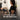 Woman uses a muscle stimulator on her back while man exercises on the floor indoors.